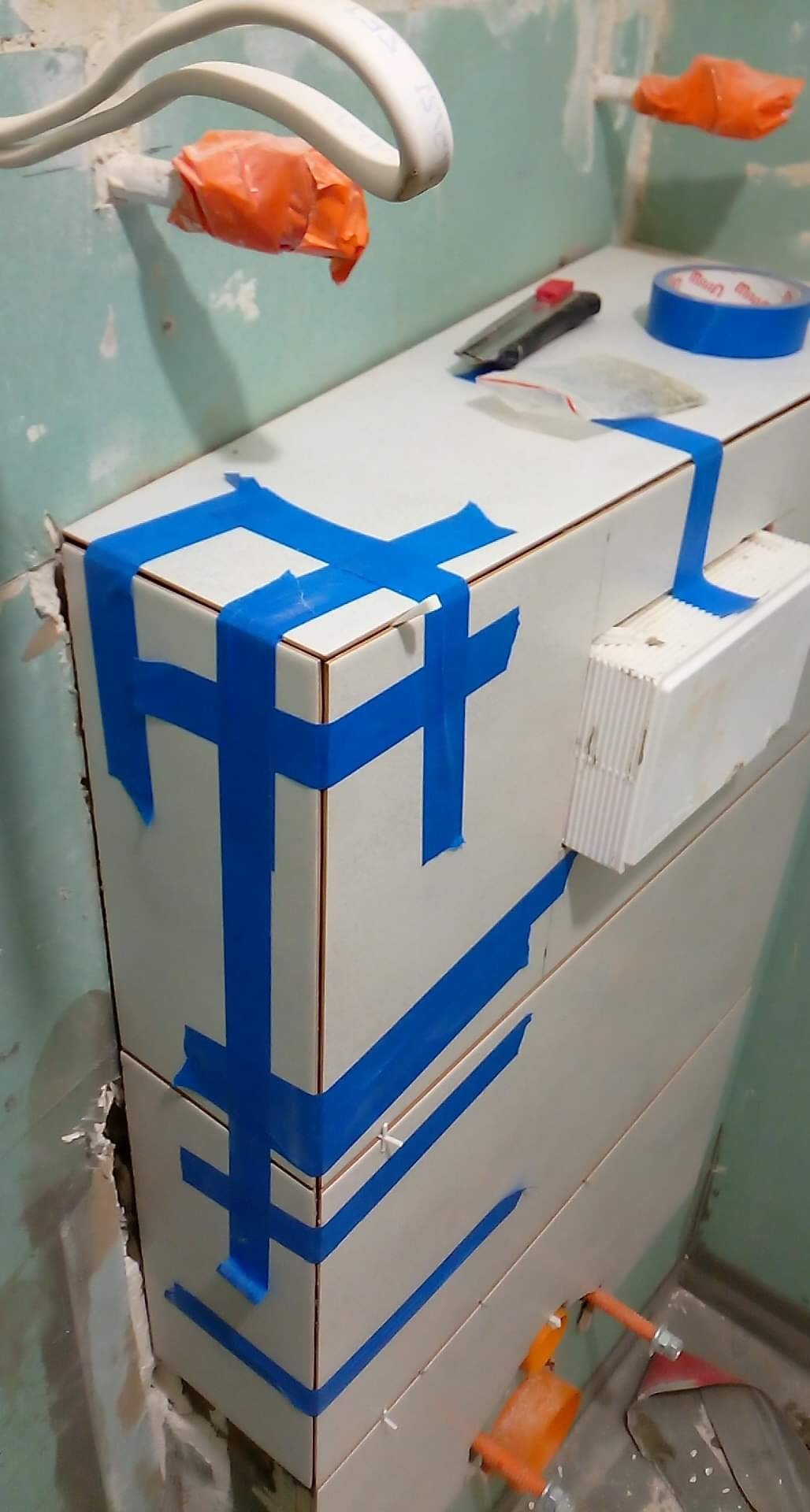 Biała zabudowa z płytek, zabezpieczona niebieską taśmą malarską, widoczne narzędzia glazurnicze i instalacja elektryczna w trakcie prac wykończeniowych.