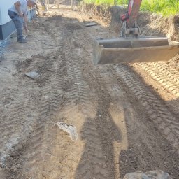 Łukasz Siemieńczuk - Czerwona koparka z łyżką wyrównuje teren przy nowo wybudowanym domu, widoczne ślady opon na ziemi, mężczyzna przygląda się pracy