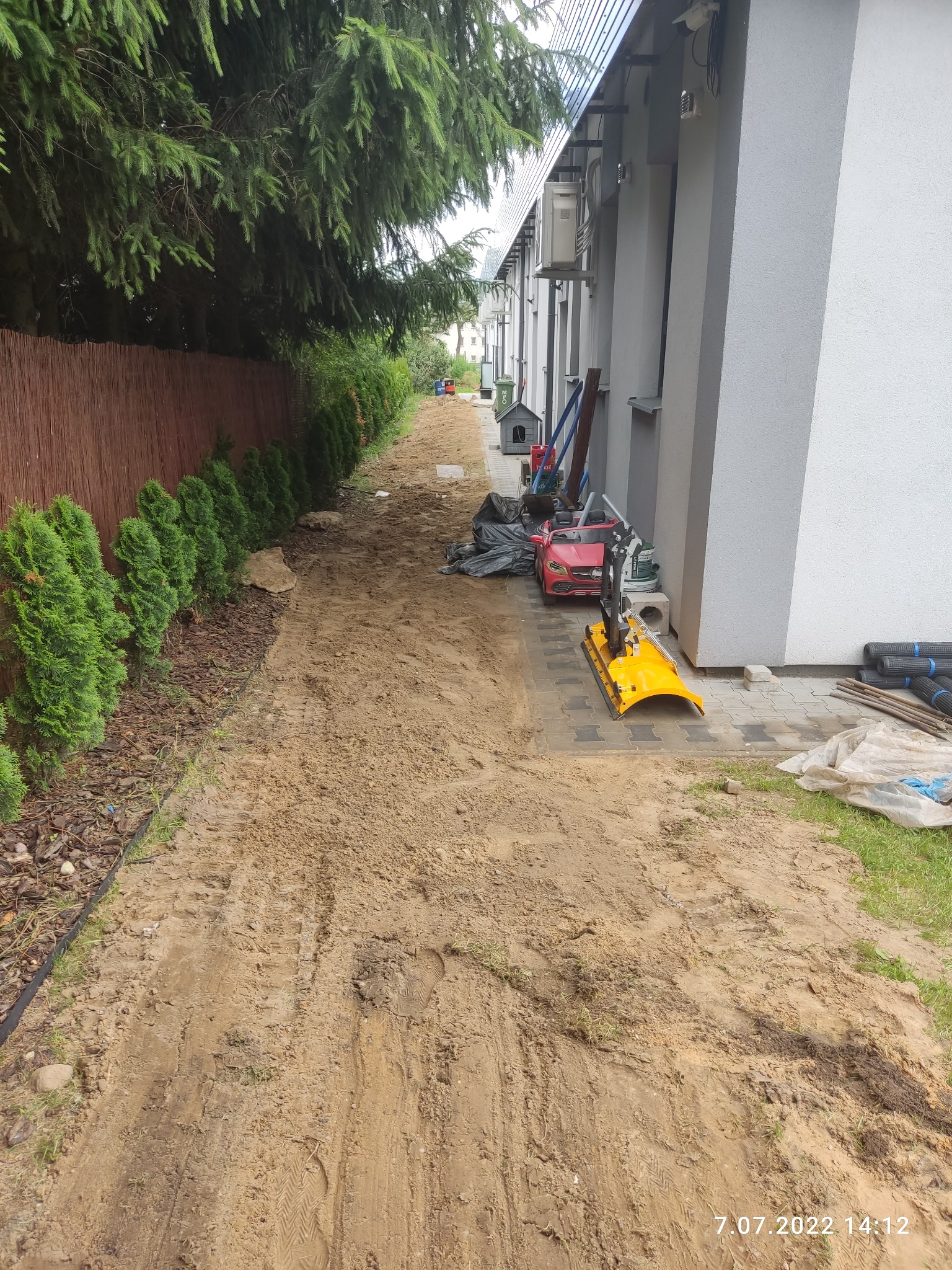 Przygotowany teren pod trawnik lub chodnik, widoczne ślady opon i butów na piasku, obok domu leżą narzędzia i materiały budowlane, w tle rząd tuj i brązowe ogrodzenie.