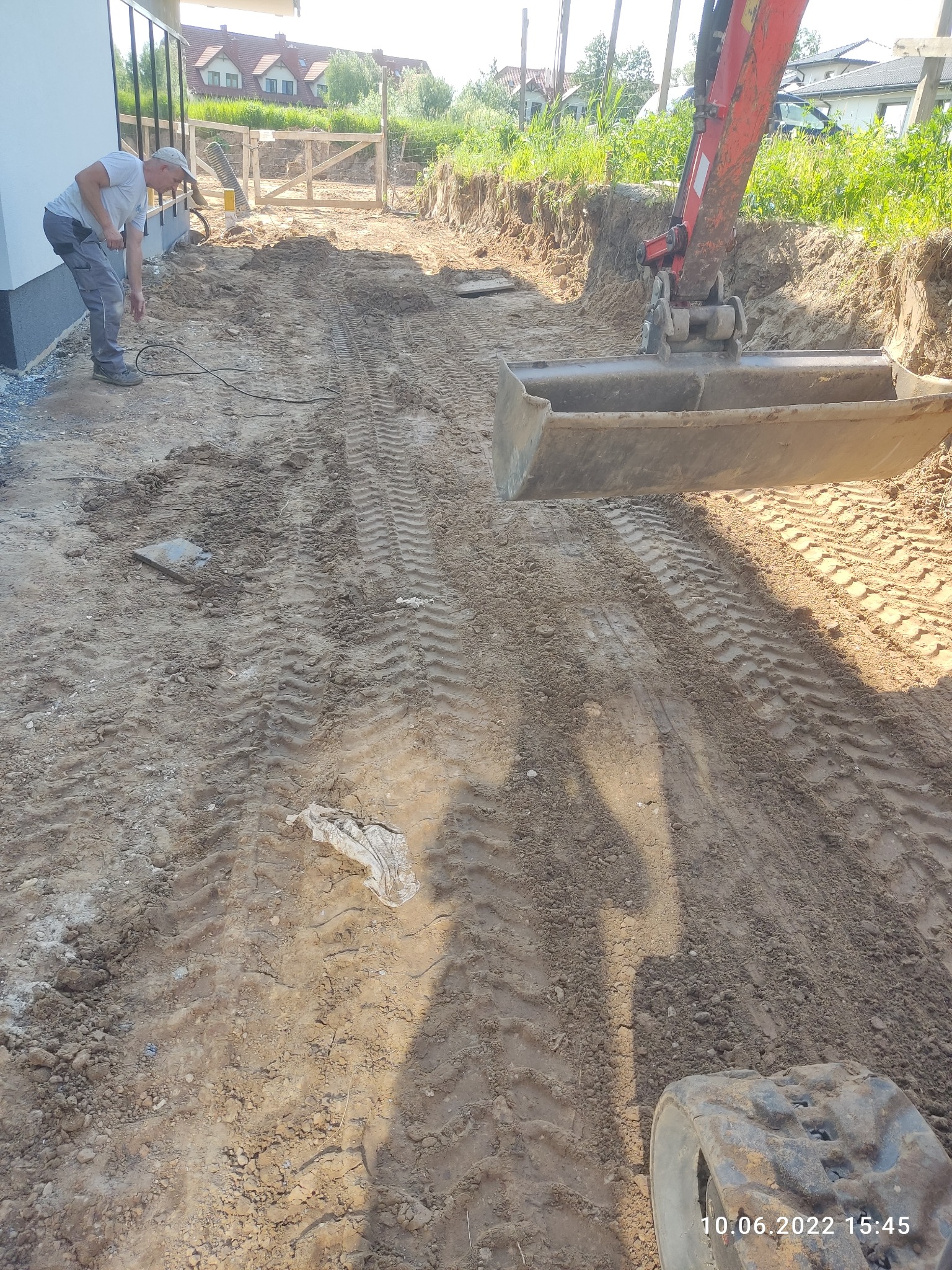 Czerwona koparka z łyżką wyrównuje teren przy nowo wybudowanym domu, widoczne ślady opon na ziemi, mężczyzna przygląda się pracy