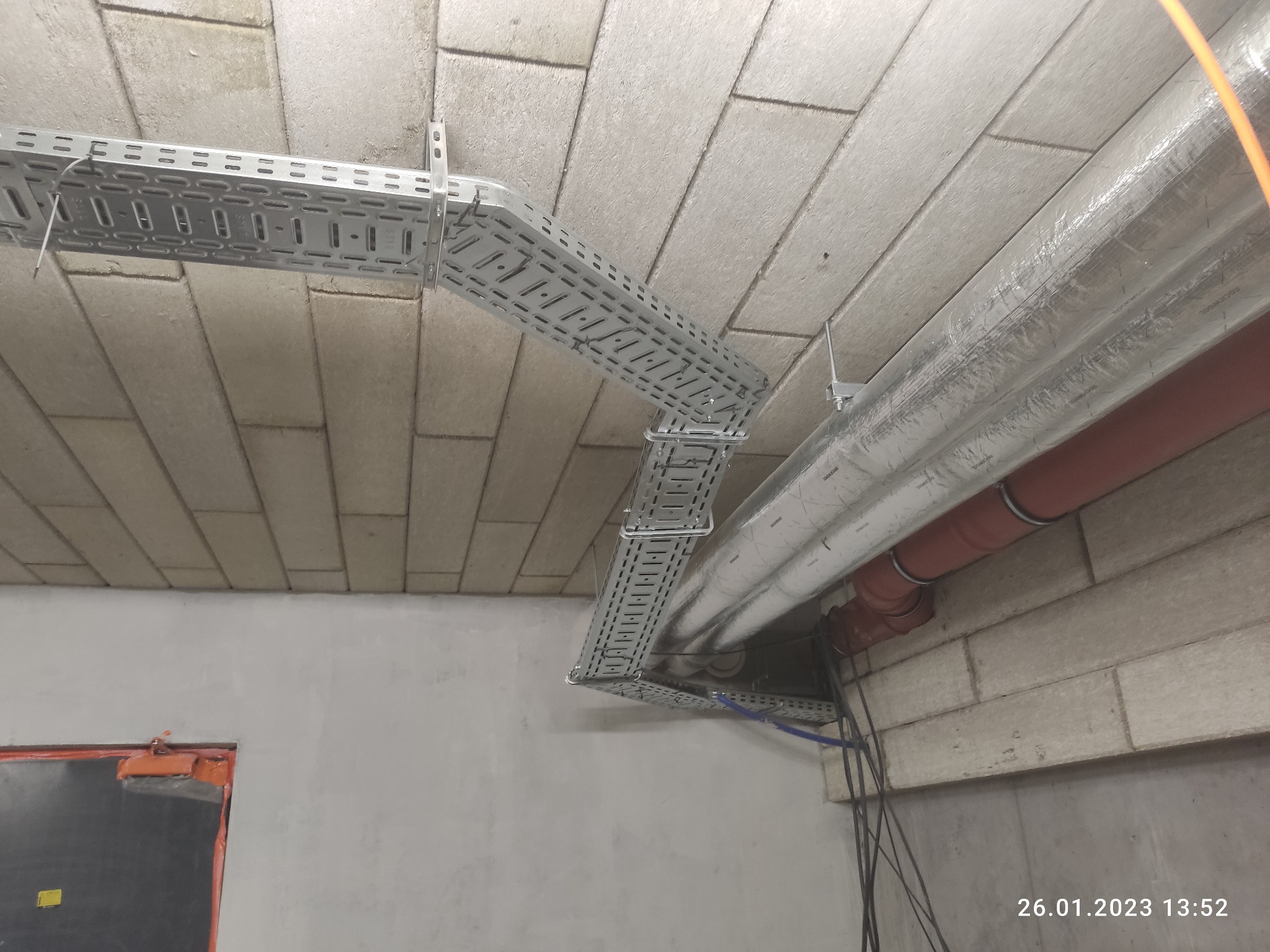 Metalowe korytko kablowe zamocowane pod sufitem z widocznymi kablami i rurami wentylacyjnymi w surowym wnętrzu.