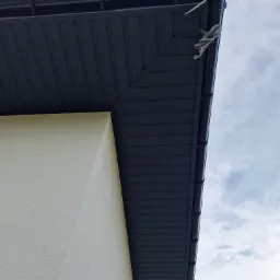 Nowoczesne, grafitowe podbitki dachowe zamontowane w Sieprawiu, widok z dołu, z fragmentem elewacji w kolorze kremowym i zwisającym kablem.