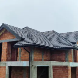 Dom w stanie surowym otwartym z zamontowaną, grafitową dachówką ceramiczną i orynnowaniem, widoczne ceglane ściany i betonowe elementy konstrukcyjne.