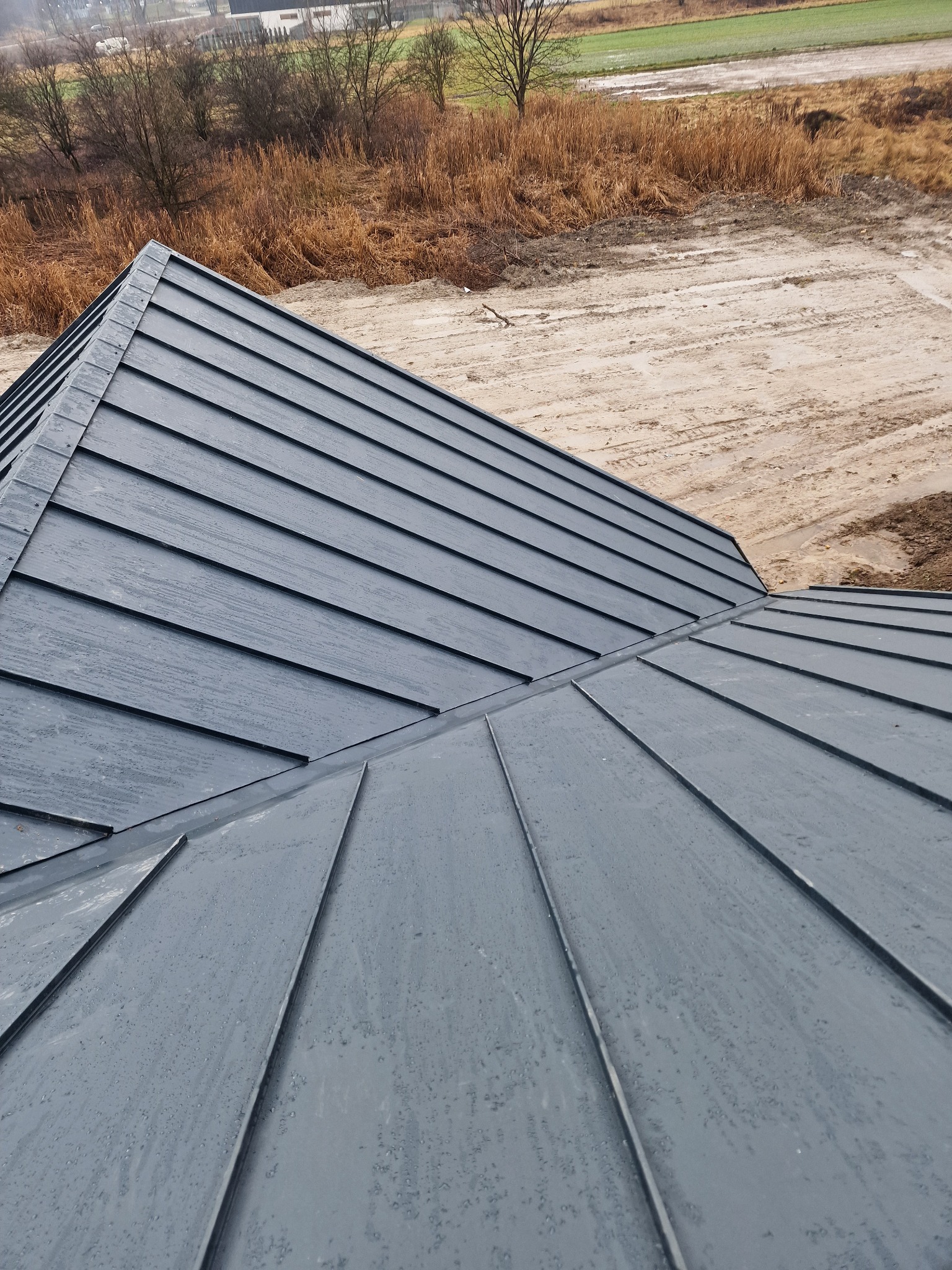 Nowy, ciemnoszary dach z blachy na tle jesiennego krajobrazu z polami i drzewami bez liści.