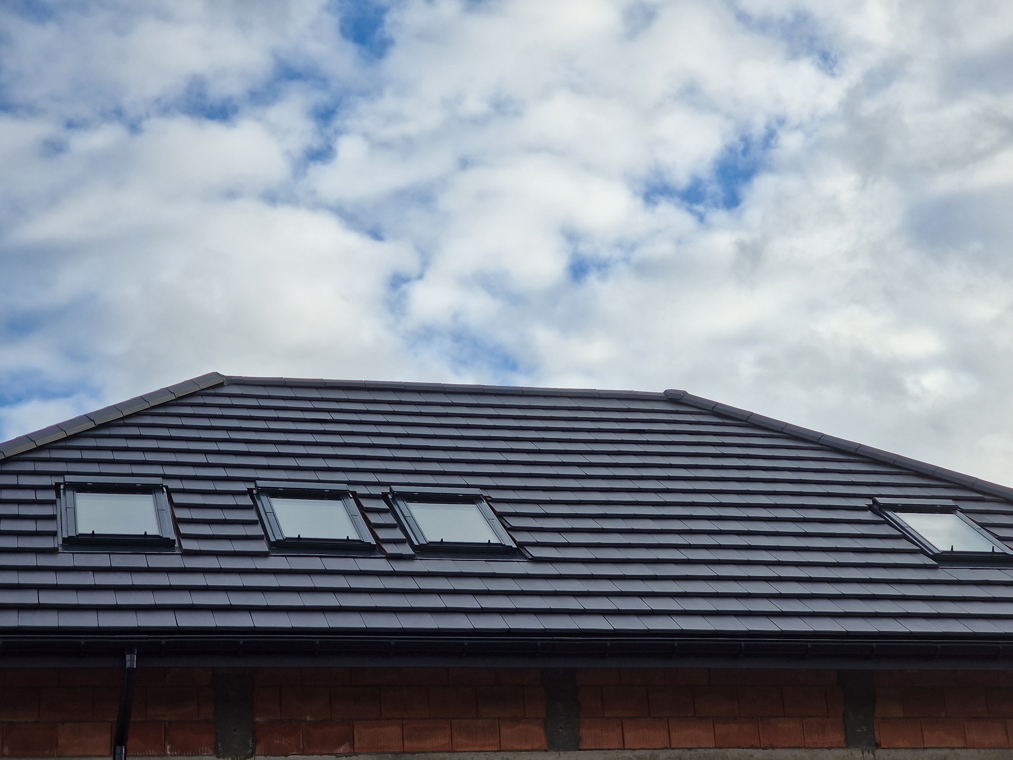 Nowy dach z ciemnej dachówki ceramicznej z trzema oknami dachowymi, widok z dołu na tle błękitnego nieba z białymi chmurami, mur z cegły widoczny w dolnej części kadru.