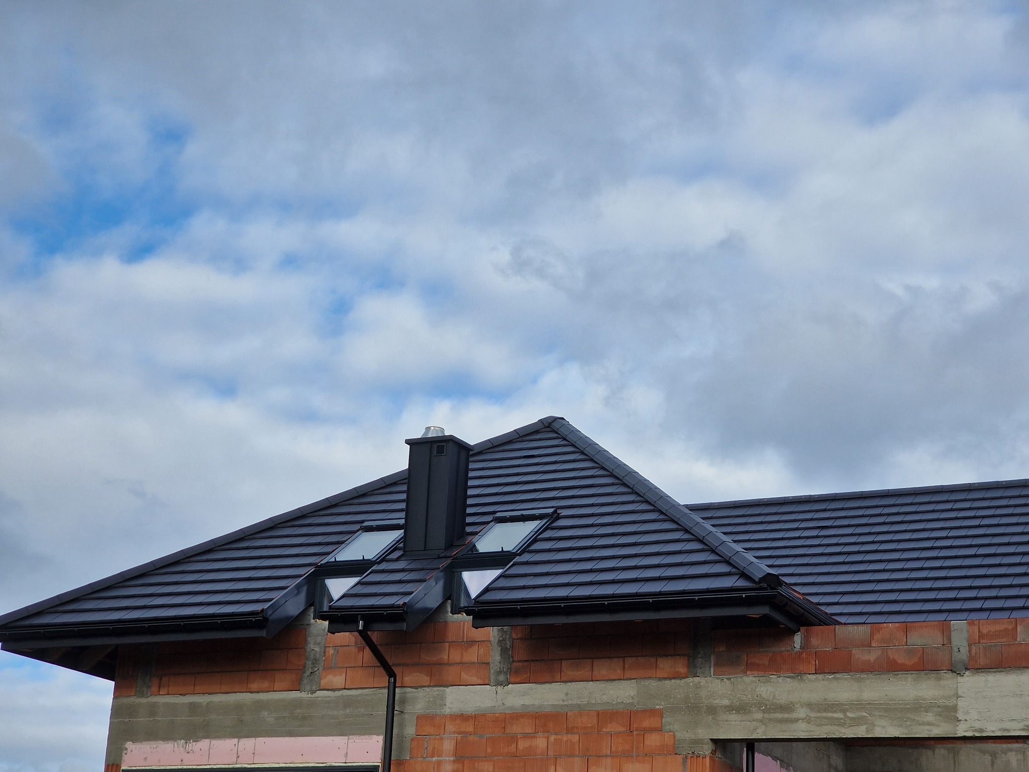 Nowy, ciemny dach z dachówką ceramiczną i oknami dachowymi na budynku w trakcie budowy, na tle pochmurnego nieba.