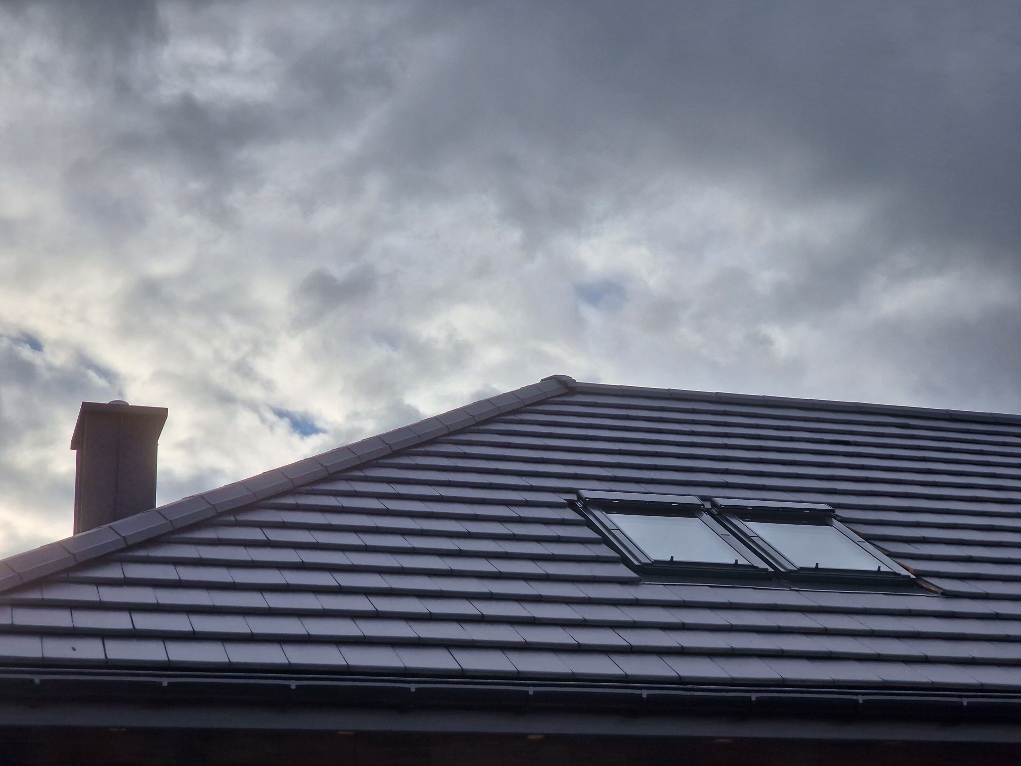 Nowy dach z ciemnej dachówki ceramicznej z dwoma oknami dachowymi i kominem na tle pochmurnego nieba.