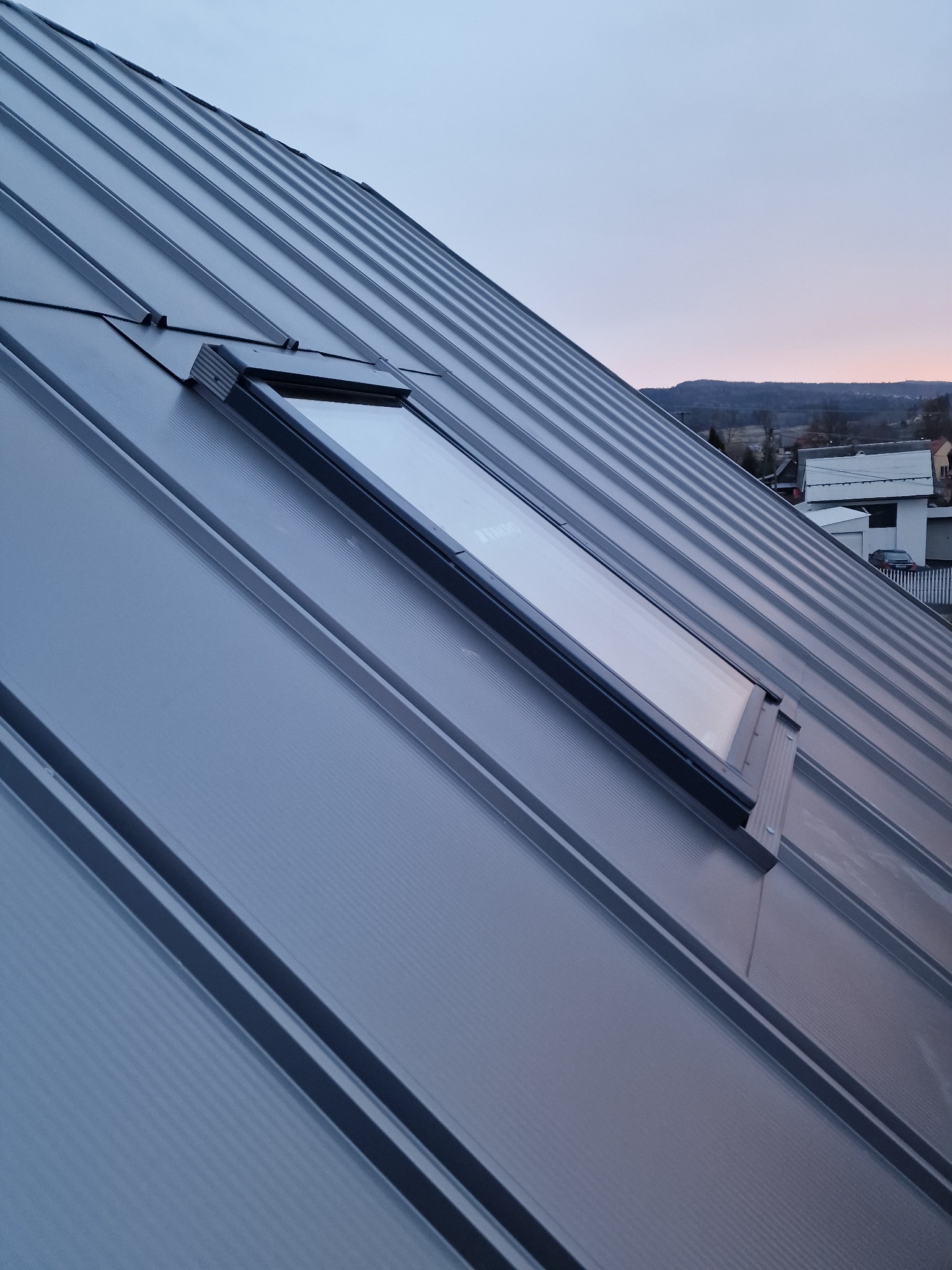Nowy, metalowy dach panelowy z oknem dachowym Fakro, widok z dołu pod kątem, w tle zabudowa jednorodzinna i zachodzące słońce.