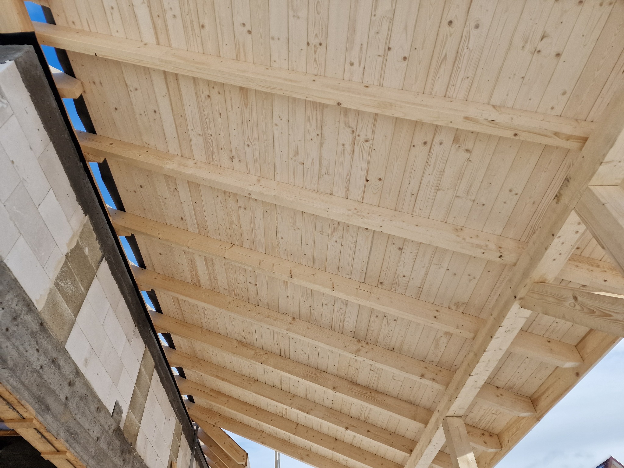 Konstrukcja drewniana dachu od wewnątrz, widoczne deski i elementy więźby dachowej, fragment ściany z betonu komórkowego w tle, widoczne prześwity nieba.