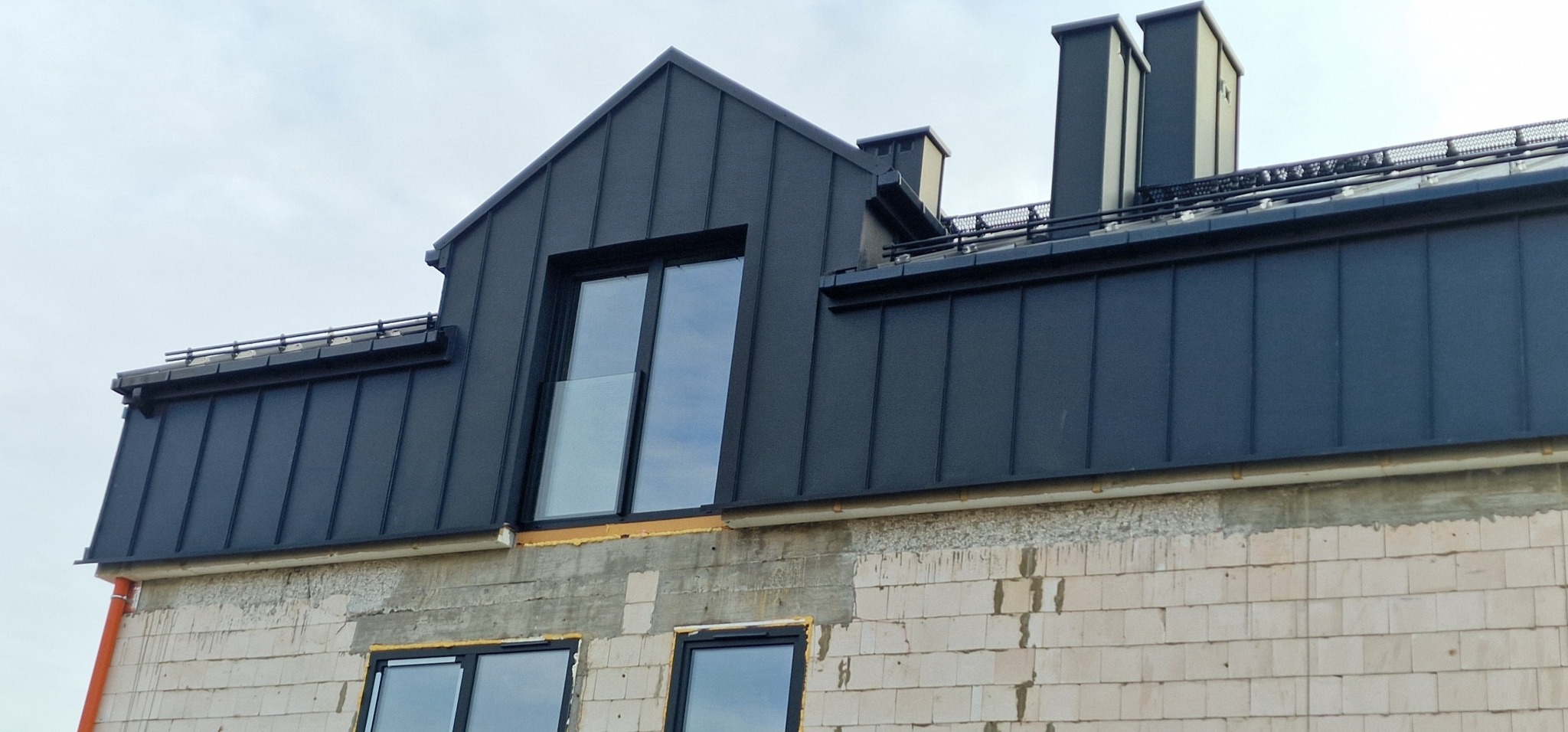 Nowoczesny, grafitowy dach z blachy na budynku w trakcie budowy. Widoczne okna dachowe i kominy. Charakterystyczne metalowe zabezpieczenia przeciwśniegowe.