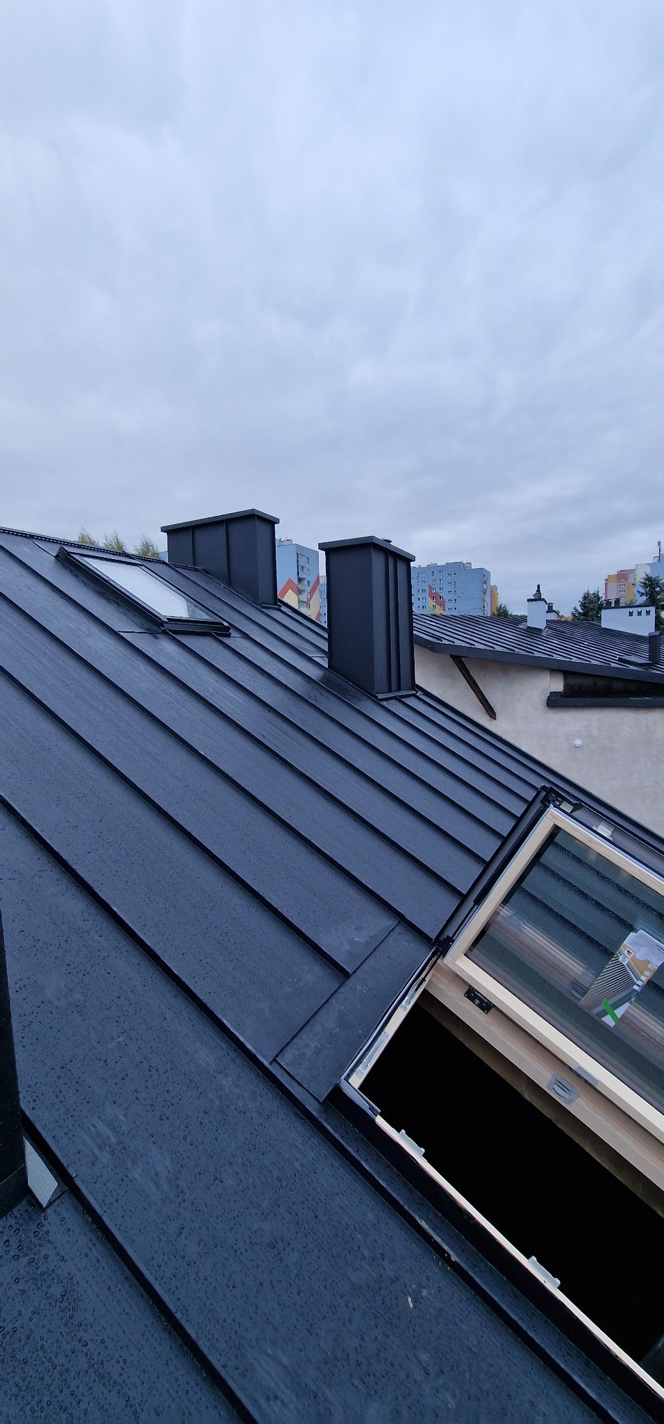 Czarny dach z blachy na budynku wielorodzinnym, widoczne kominy i okno dachowe w trakcie montażu. Na dachu widoczne krople deszczu, pochmurne niebo w tle.