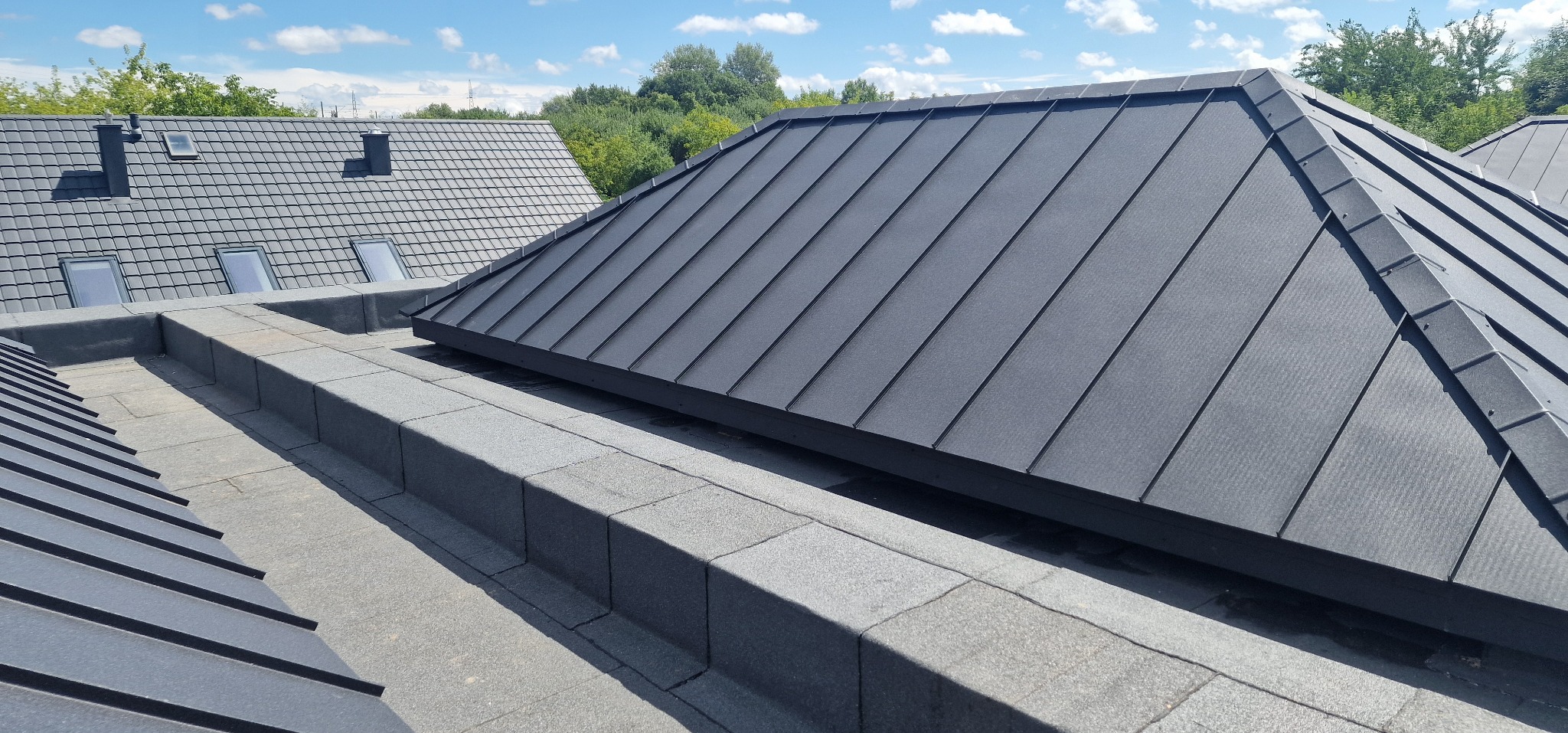 Szary dach z blachy na budynku z widocznymi elementami obróbki blacharskiej i fragmentem dachu z dachówki w tle, w słoneczny dzień.