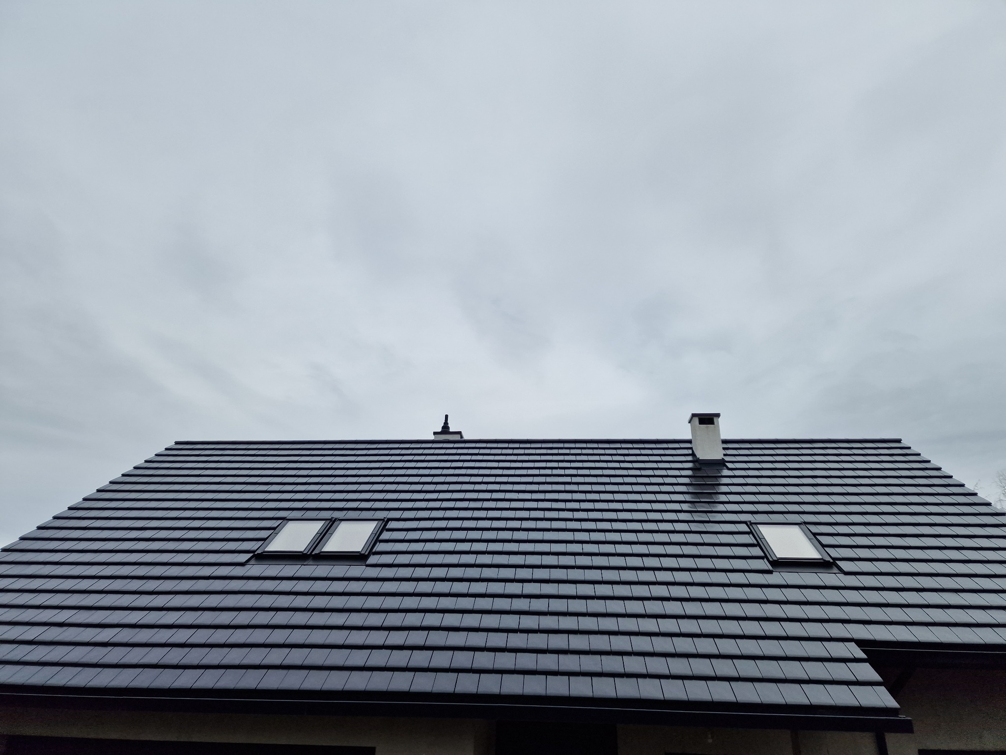 Ciemna dachówka ceramiczna na skośnym dachu z dwoma oknami dachowymi i kominem na tle pochmurnego nieba. Widok z dołu.