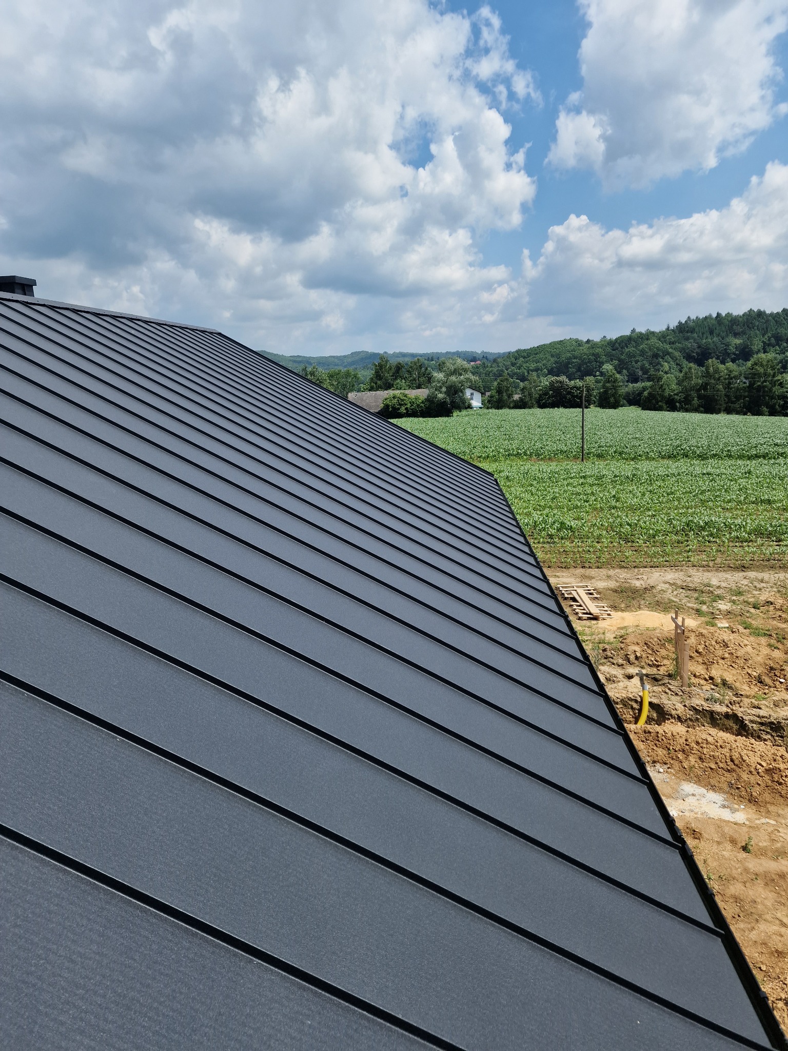 Nowy, grafitowy dach z blachy na tle zielonego pola uprawnego i błękitnego nieba z chmurami.