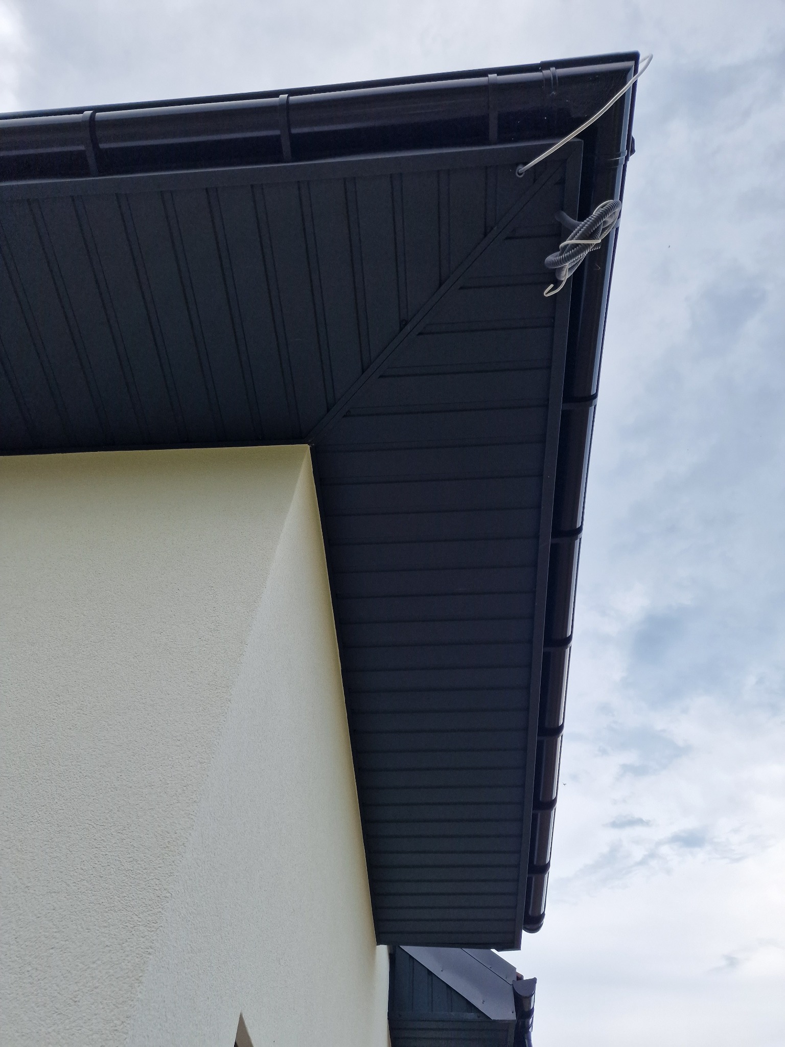 Nowoczesne, grafitowe podbitki dachowe zamontowane w Sieprawiu, widok z dołu, z fragmentem elewacji w kolorze kremowym i zwisającym kablem.