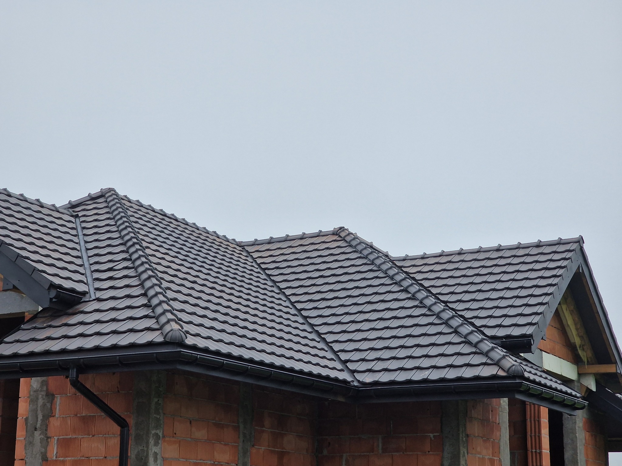 Nowy dach z brązowej dachówki ceramicznej na surowym budynku z czerwonej cegły, widoczne orynnowanie i niezakończone prace budowlane.