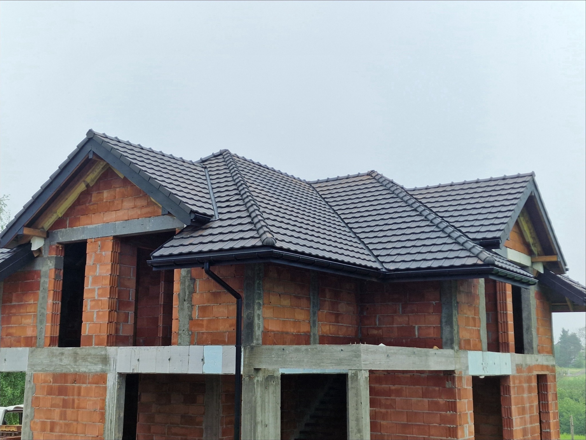 Dom w stanie surowym otwartym z zamontowaną, grafitową dachówką ceramiczną i orynnowaniem, widoczne ceglane ściany i betonowe elementy konstrukcyjne.