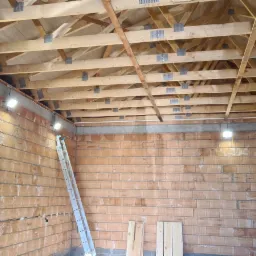 Wnętrze w trakcie budowy z widoczną konstrukcją drewnianą dachu, murowanymi ścianami z pustaków ceramicznych, aluminiową drabiną opartą o ścianę i zamontowanymi lampami LED na ścianach.
