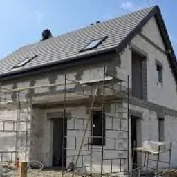 Niedokończony, dwukondygnacyjny budynek z szarej cegły, z szarym dachem i oknami dachowymi, otoczony rusztowaniem, w trakcie prac wykończeniowych.