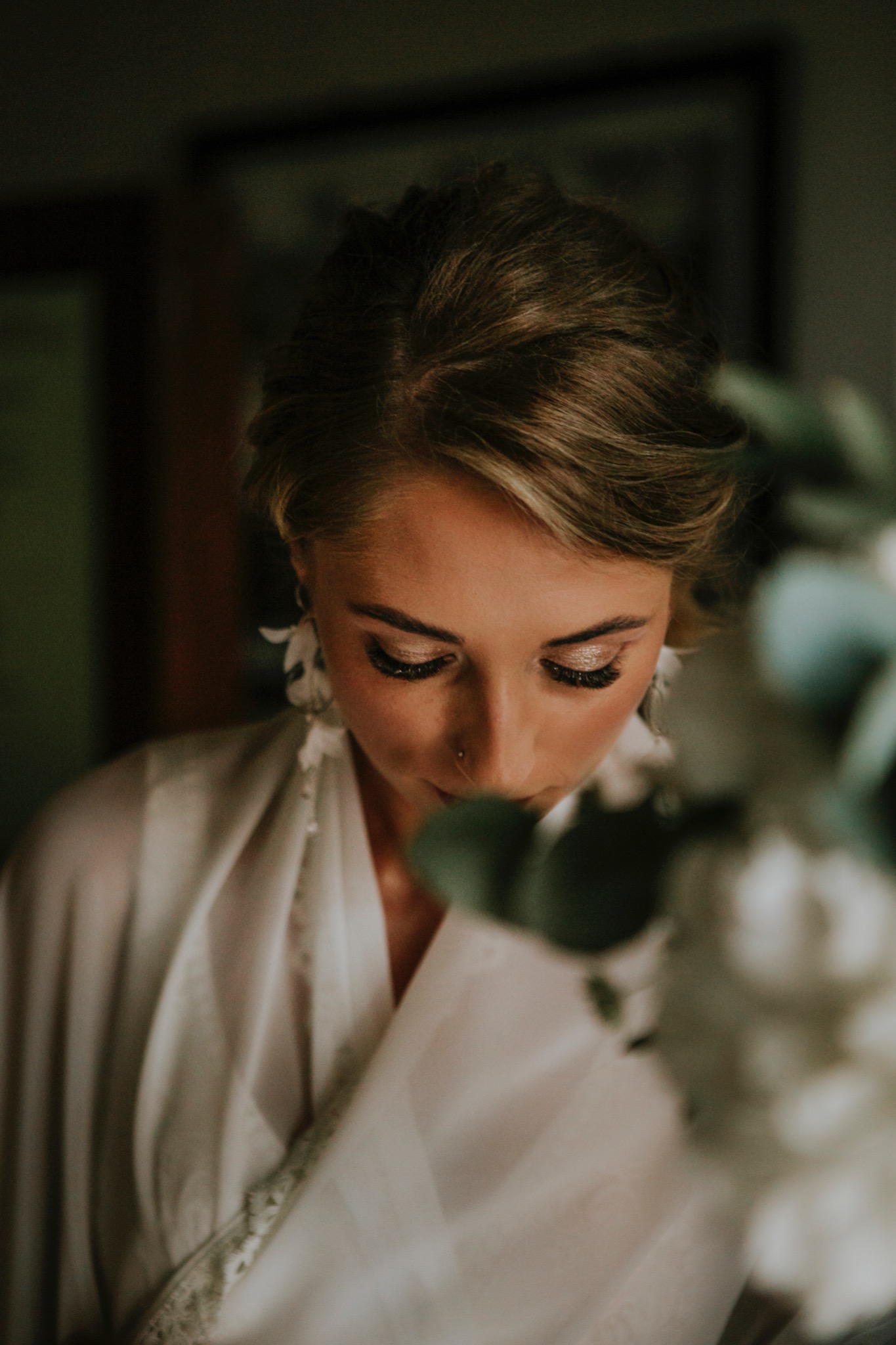 Portret kobiety z delikatnym makijażem i kwiatowymi kolczykami, ubrana w zwiewną, białą szatę, spoglądająca w dół wśród zieleni.