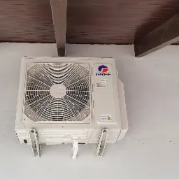 Biała jednostka zewnętrzna klimatyzacji marki Gree zamontowana na jasnej ścianie pod drewnianą konstrukcją dachu.