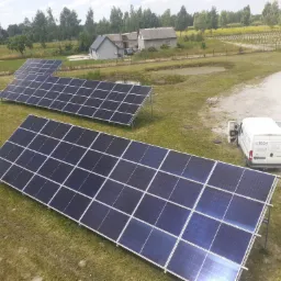 Widok z góry na instalację fotowoltaiczną na trawiastym terenie, z białym busem serwisowym ELTECH w tle i budynkami mieszkalnymi w oddali.