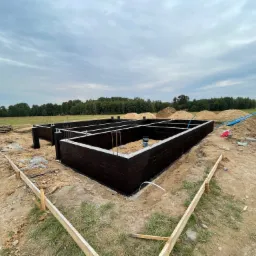 Zabezpieczone fundamenty domu jednorodzinnego, widoczne zbrojenia i izolacja przeciwwilgociowa, w tle teren budowy z hałdami piasku i fragmentem lasu.