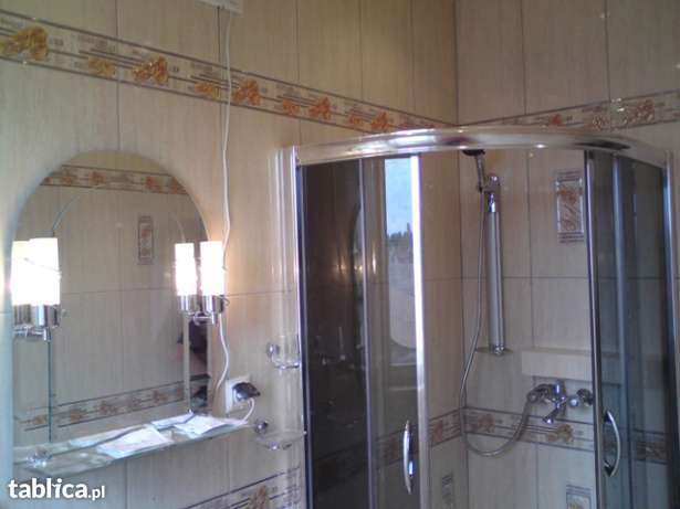 Wykończona łazienka z kabiną prysznicową, lustrem z oświetleniem i beżowymi płytkami z dekoracyjnym pasem, widoczne gniazdko elektryczne przy lustrze.