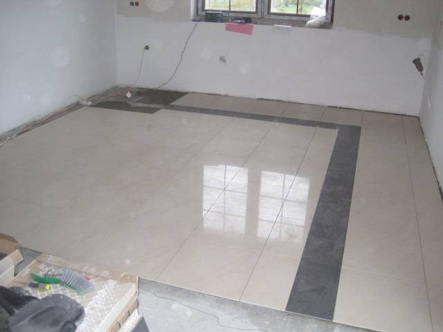 Układanie beżowych płytek podłogowych z ciemnoszarym obramowaniem w jasnym pomieszczeniu w trakcie remontu, widoczne narzędzia i niedokończone fragmenty podłogi.