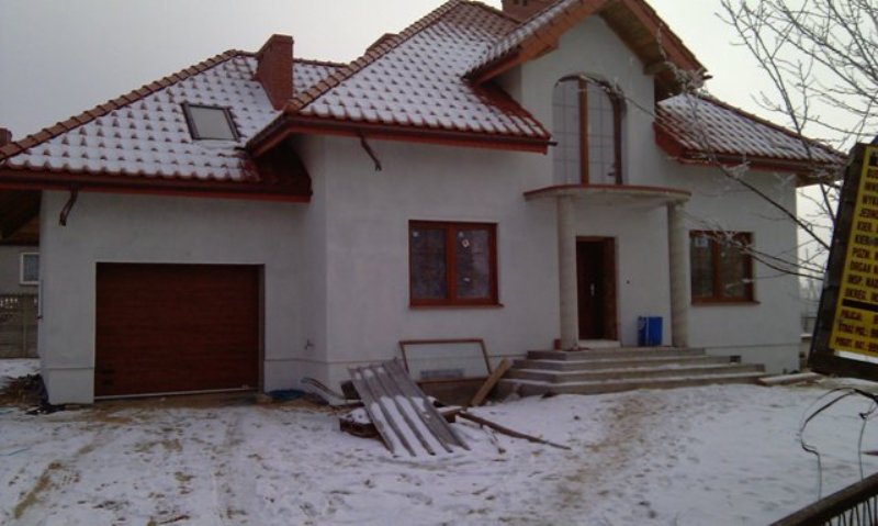 Nowo wybudowany, otynkowany dom murowany z brązową bramą garażową i dachem pokrytym czerwoną dachówką, częściowo zaśnieżony, w zimowej scenerii.