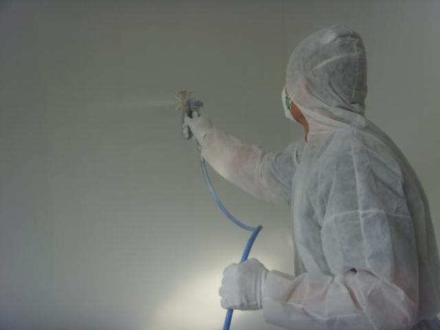 Pracownik w kombinezonie ochronnym maluje ścianę metodą natryskową, widoczny pistolet lakierniczy z niebieskim przewodem.