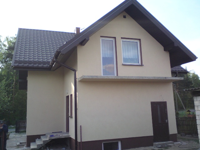 Dwukondygnacyjny dom z beżową elewacją, ciemnobrązowym dachem z dachówki, białymi oknami z brązowymi ramami, widoczny z zewnątrz, z elementami docieplenia.