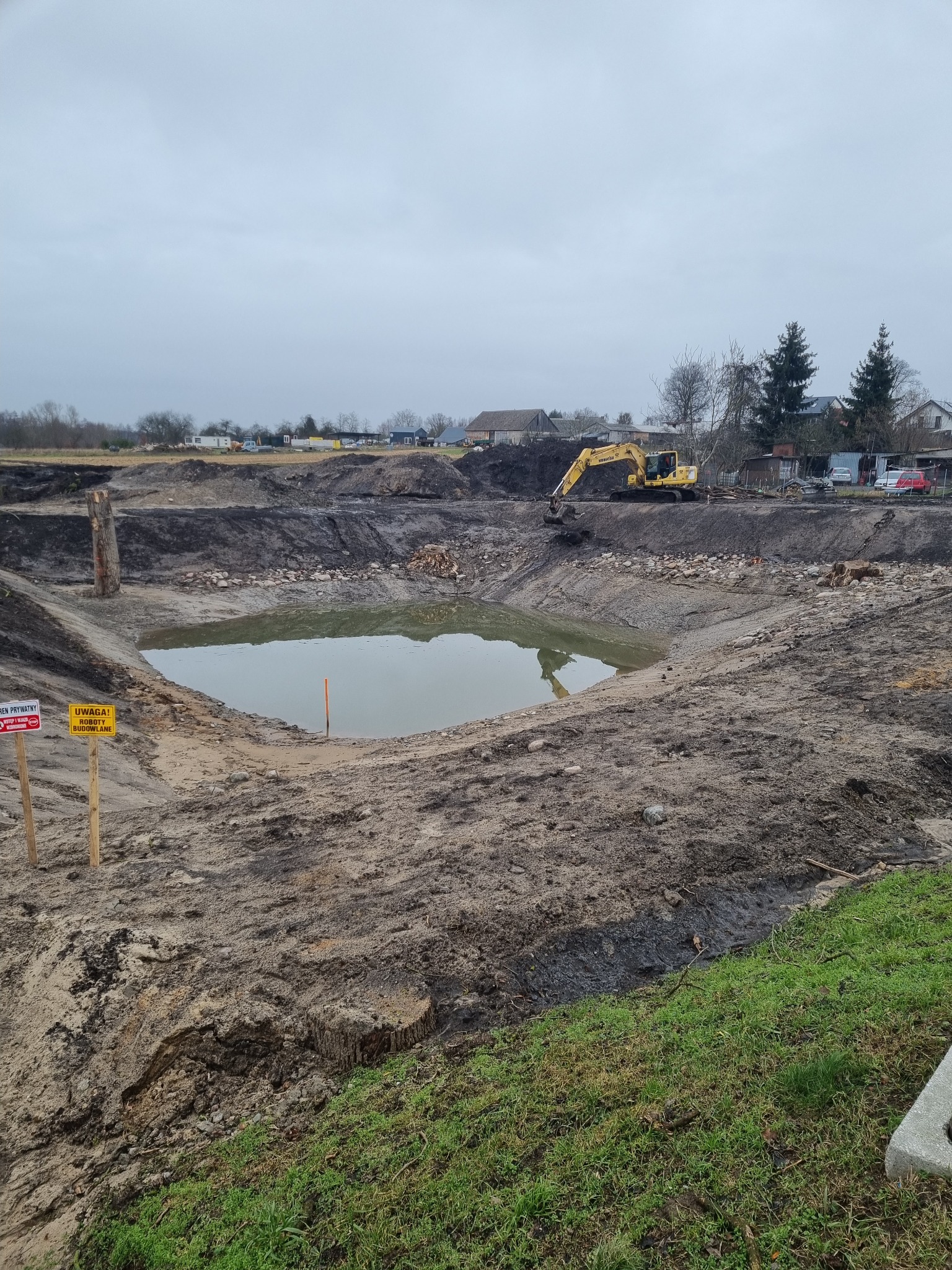 Żółta koparka na brzegu wykopanego stawu wypełnionego wodą, tablice informacyjne o robotach budowlanych, pochmurne niebo w tle.