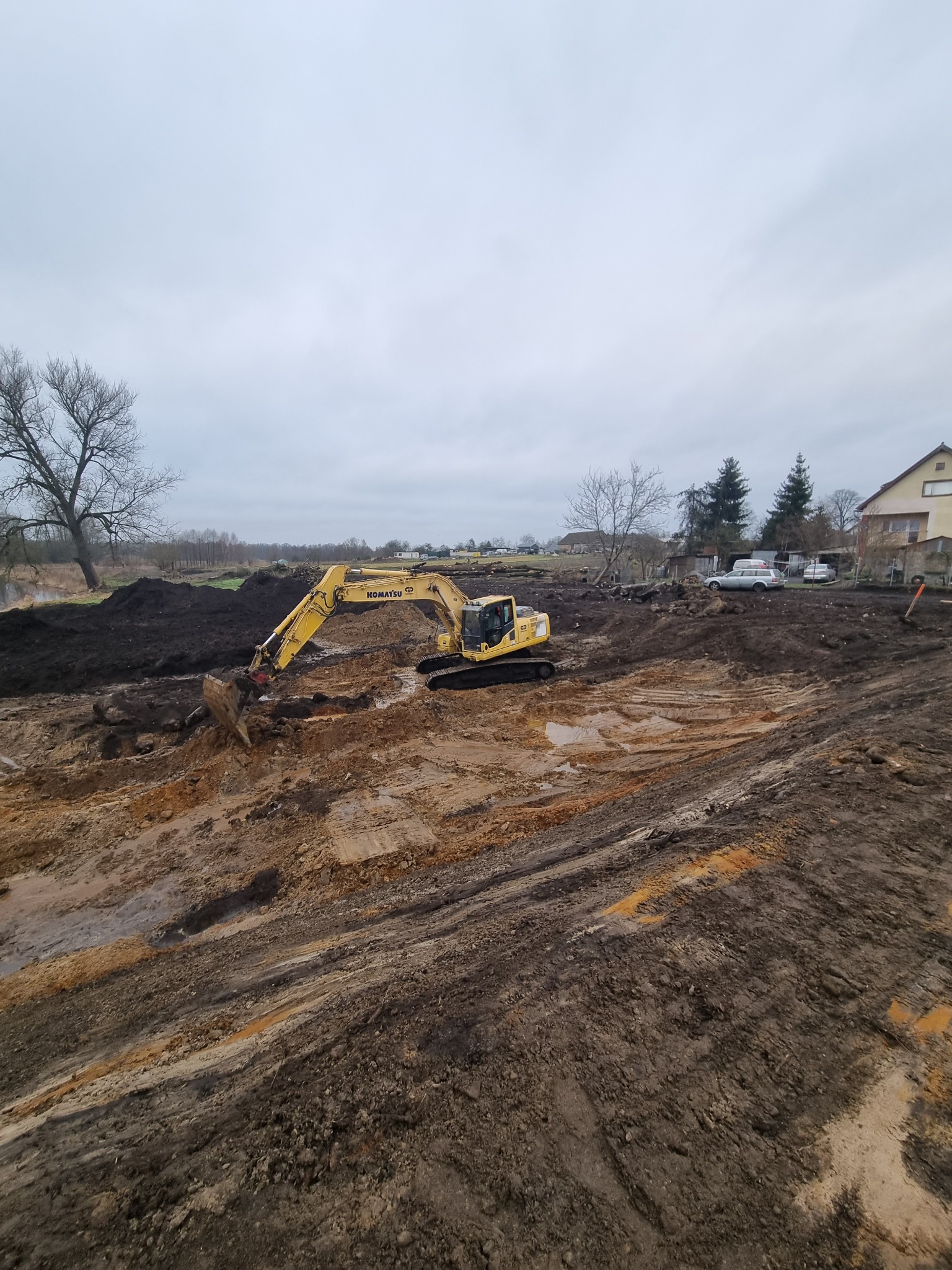 Żółta koparka Komatsu w trakcie prac ziemnych na podmokłym terenie, widoczne ślady opon w błocie i hałdy ziemi, w tle zabudowania i pochmurne niebo.