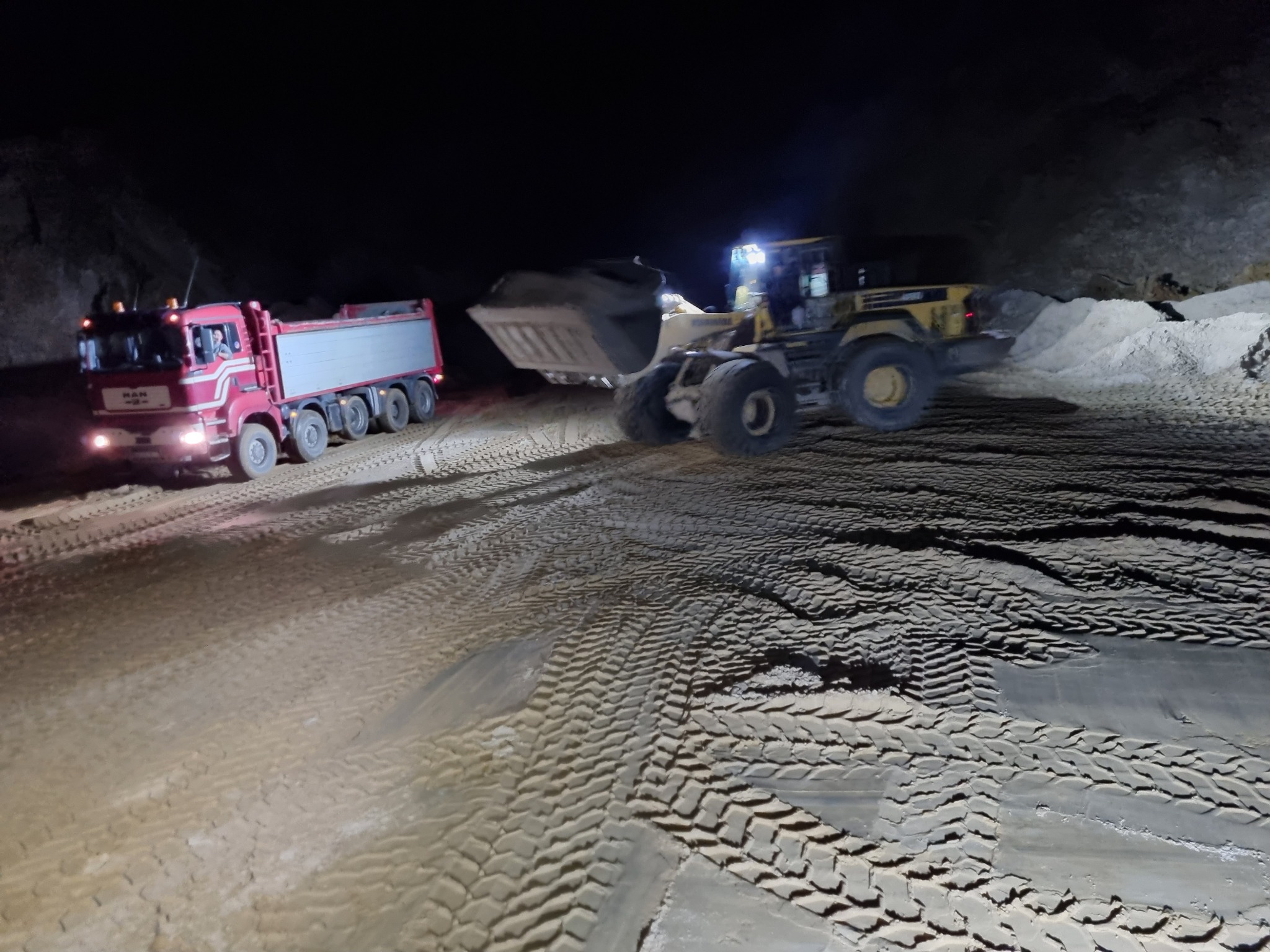 Żółta ładowarka kołowa nabiera materiał do łyżki, obok stoi czerwona ciężarówka MAN, widoczne ślady opon na ziemi, nocna sceneria.