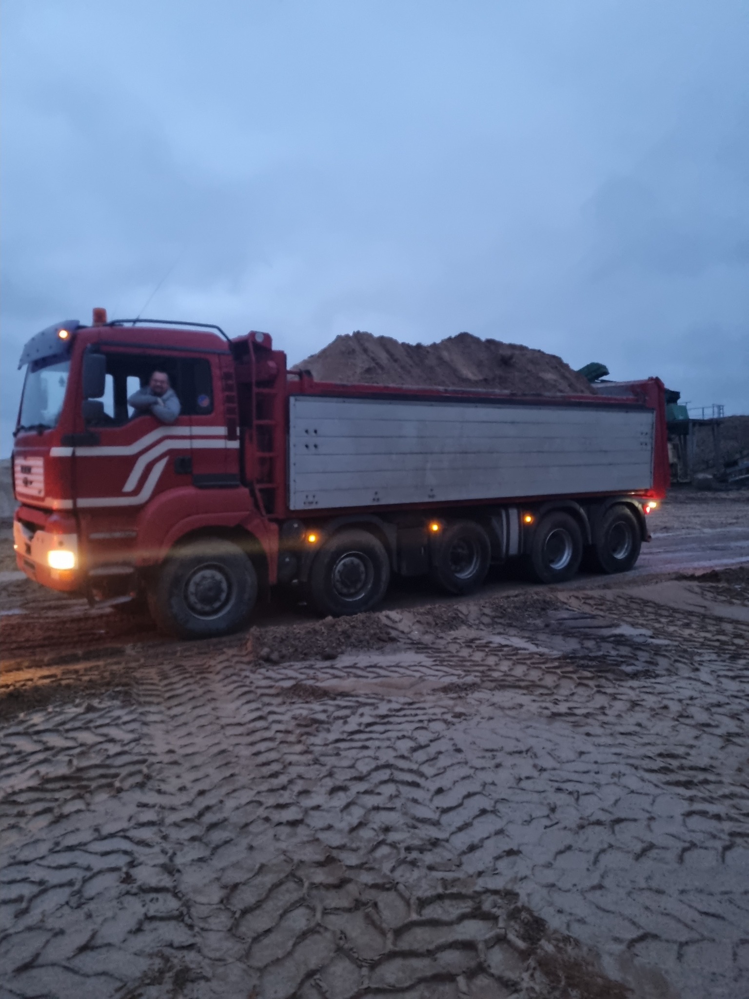 Czerwona ciężarówka z załadowaną piaskiem skrzynią stoi na ubitym podłożu, widoczny kierowca w kabinie, oświetlenie zmierzchowe.