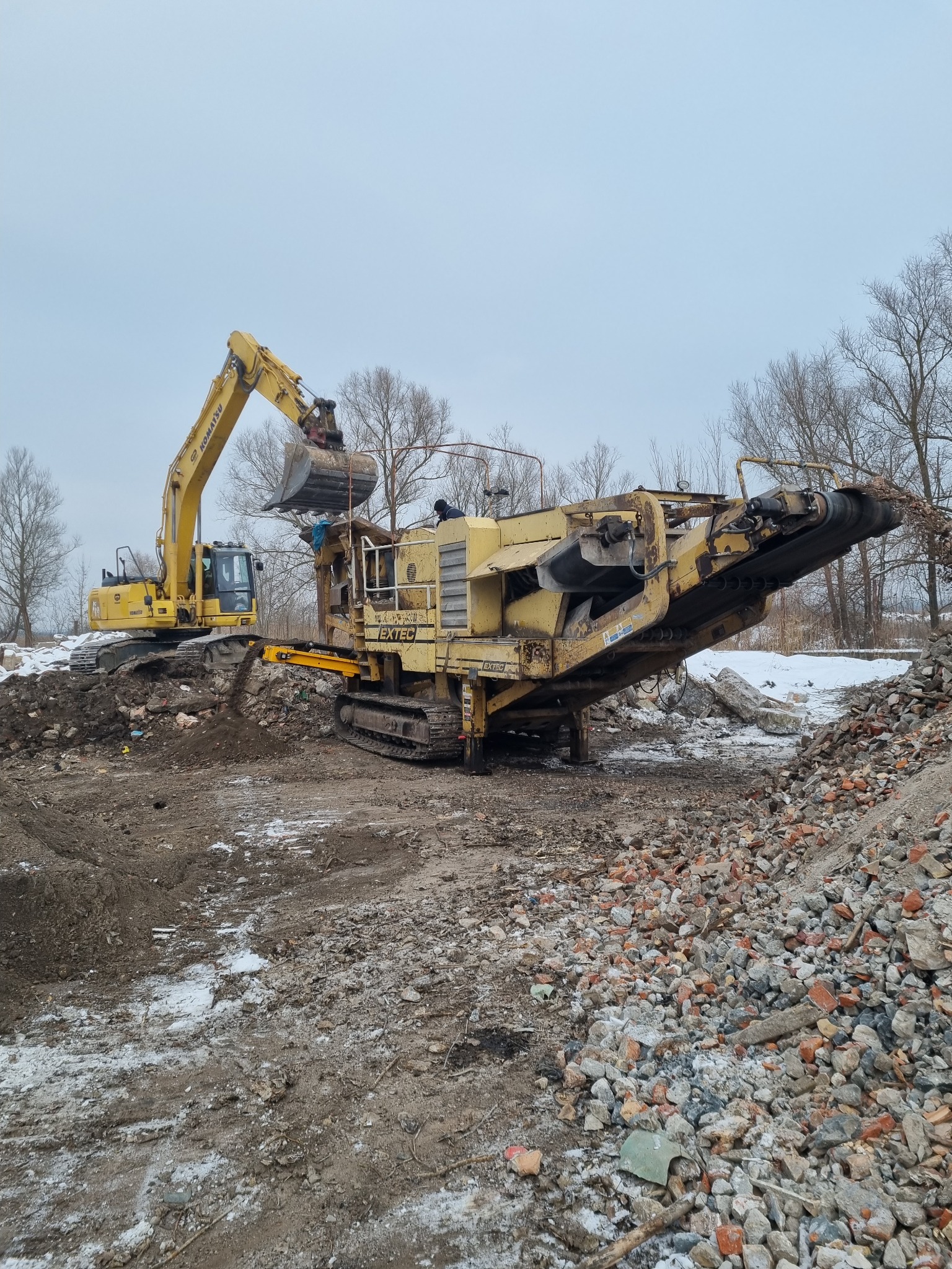 Żółta koparka Komatsu podnosi gruz i wrzuca go do żółtej kruszarki EXTEC na tle drzew bez liści i szarego nieba; teren częściowo pokryty śniegiem.