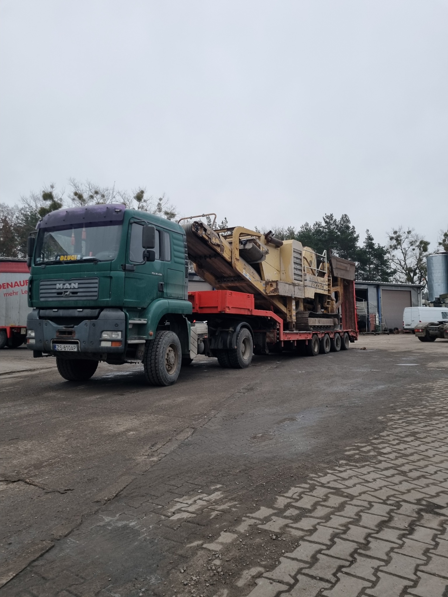 Zielony ciągnik MAN z naczepą transportuje żółtą maszynę do kruszenia kamieni EXTEC na placu z kostki brukowej i ziemi.