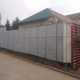 Wznoszenie żelbetowej ściany oporowej przy budowie domu, widoczne szalunki i zbrojenia, w tle budynek w trakcie budowy z drewnianą konstrukcją dachu.