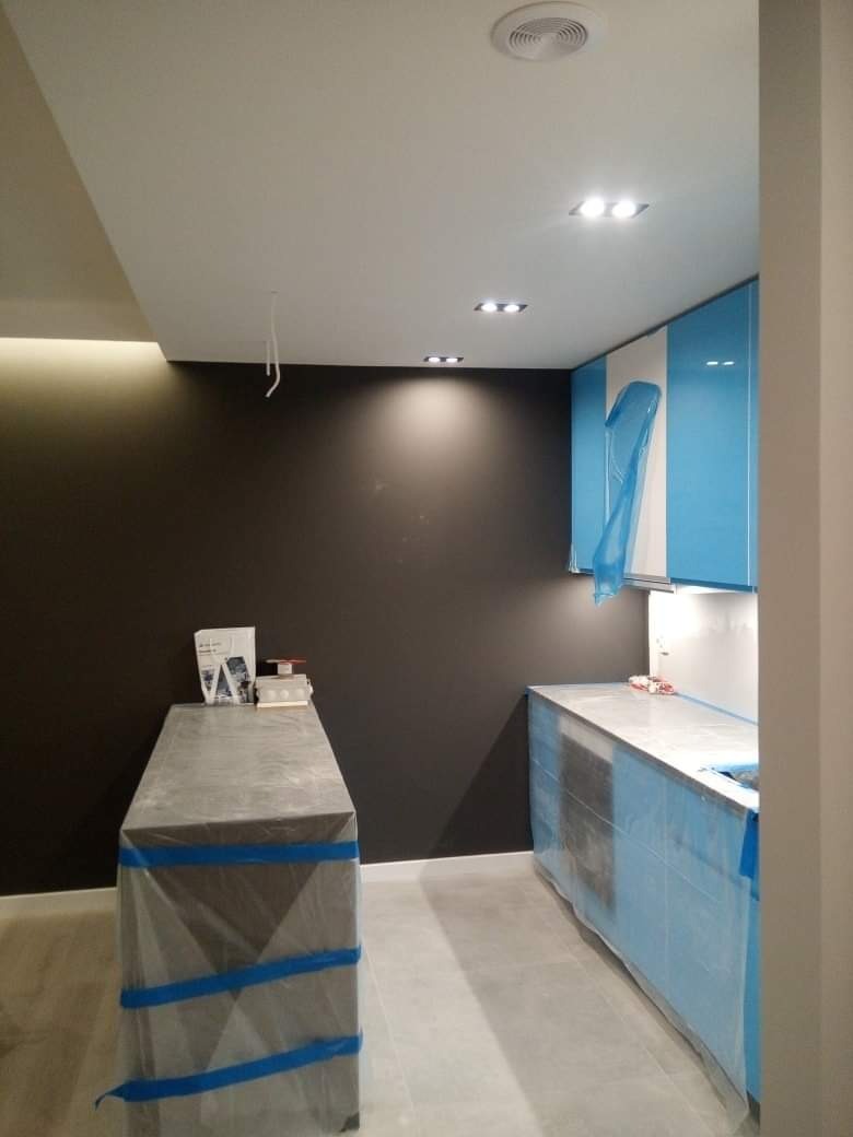 Przygotowanie kuchni do prac wykończeniowych: meble i blaty zabezpieczone folią malarską, ściana pomalowana na ciemny kolor, widoczne oświetlenie sufitowe.