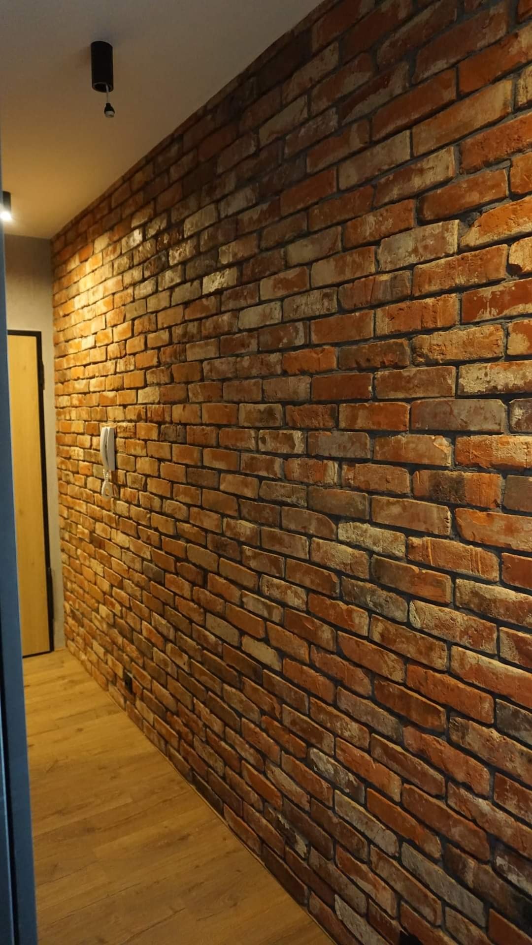 Ściana z cegły w korytarzu z drewnianą podłogą, widoczny biały telefon na ścianie i czarne oświetlenie sufitowe.