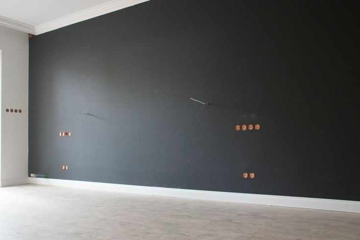 Świeżo pomalowana na czarno ściana z widocznymi puszkami instalacyjnymi i fragmentem rurki wystającej ze ściany, biała listwa przypodłogowa i jasnoszara podłoga.