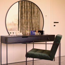 Mutacja Weronika Cwanek - Toaletka z okrągłym lustrem, czarnym blatem i zielonym, tapicerowanym krzesłem na metalowym stelażu, na blacie kosmetyki i świece.