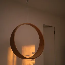 Okrągła, drewniana huśtawka wisząca na metalowym łańcuchu w minimalistycznym wnętrzu, oświetlona ciepłym światłem zachodzącego słońca, rzucającym cień rośliny na ścianę.