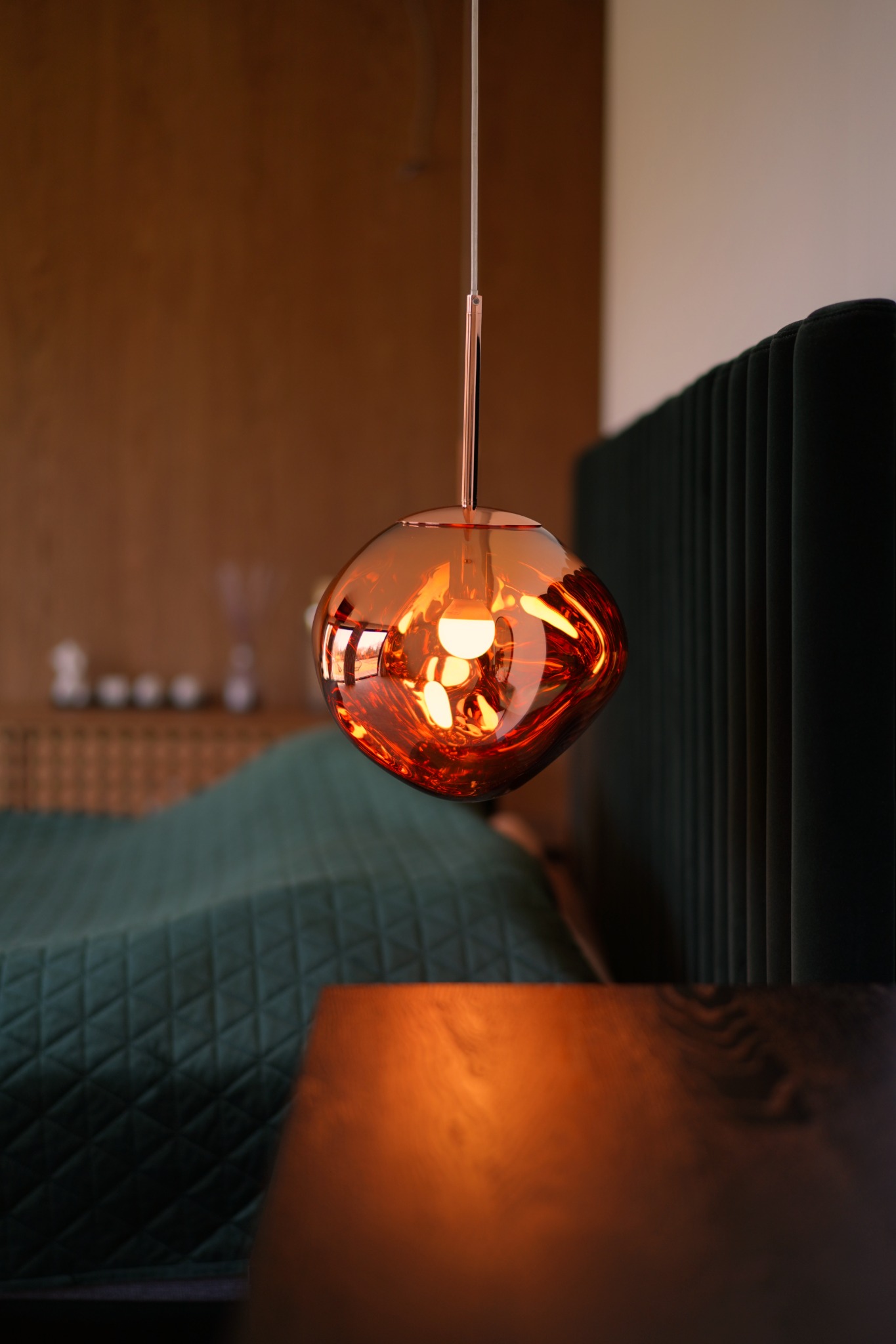 Szklana, bursztynowa lampa wisząca o nieregularnym kształcie w sypialni z zielonym zagłówkiem łóżka i drewnianą boazerią.
