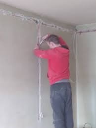 Elektryk na drabinie, mocujący przewody elektryczne do ściany w trakcie prac instalacyjnych. Widoczne kable prowadzone wzdłuż krawędzi ściany i sufitu, przygotowane do dalszego podłączenia.