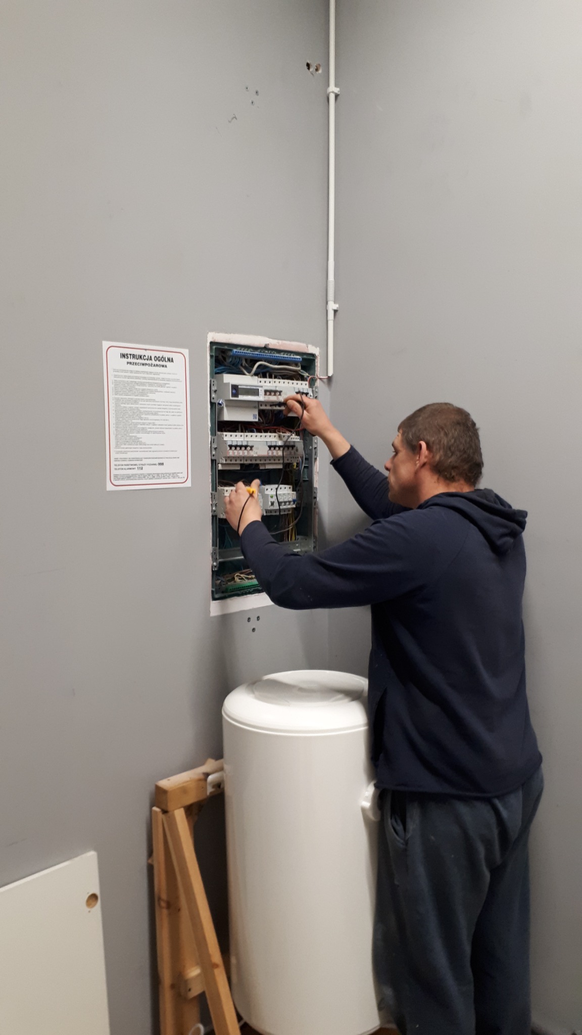 Elektryk mierzy napięcie w rozdzielnicy elektrycznej zamontowanej na szarej ścianie, obok wisi instrukcja przeciwpożarowa, a pod nią stoi biały bojler.