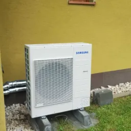 Zewnętrzna jednostka pompy ciepła marki Samsung, umieszczona na betonowych wspornikach przy żółtej ścianie budynku z małym oknem, otoczona białymi kamieniami i trawnikiem.