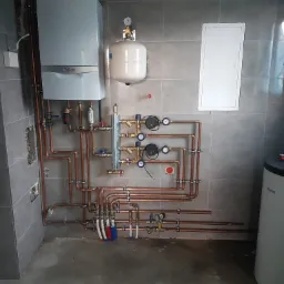 Nowoczesny piec gazowy Vaillant z rozbudowaną instalacją hydrauliczną z miedzianych rur, zbiornikiem wyr&oacute;wnawczym i pompami obiegowymi na tle szarej, kafelkowanej ściany w surowym pomieszczeniu.