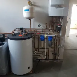 Biały, wiszący piec gazowy Vaillant z podłączonymi miedzianymi rurami, zbiornik wyrównawczy, zmiękczacz wody Water Technic i filtry wody w surowym wnętrzu budynku w trakcie wykańczania.